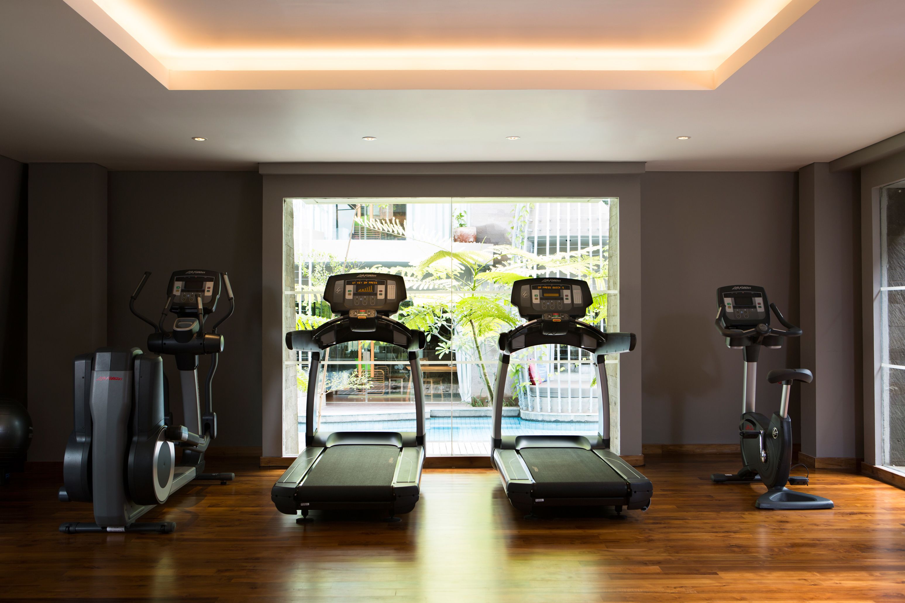 Ein Fitnessraum mit Laufbändern und Crosstrainern, große Fenster mit Blick auf einen Pool und Grünflächen.