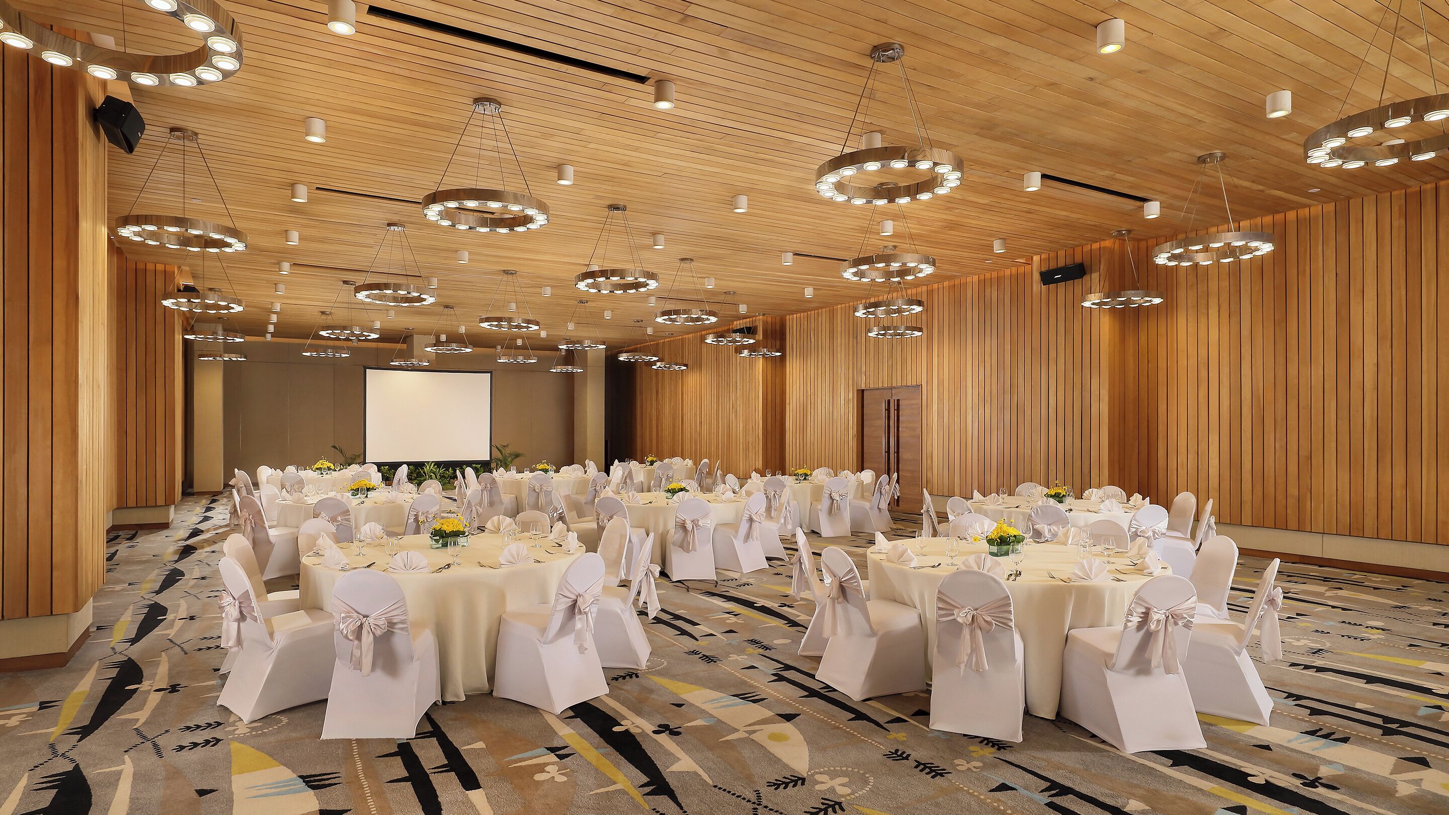 A large banquet hall with round tables, white chairs, and a projector screen.