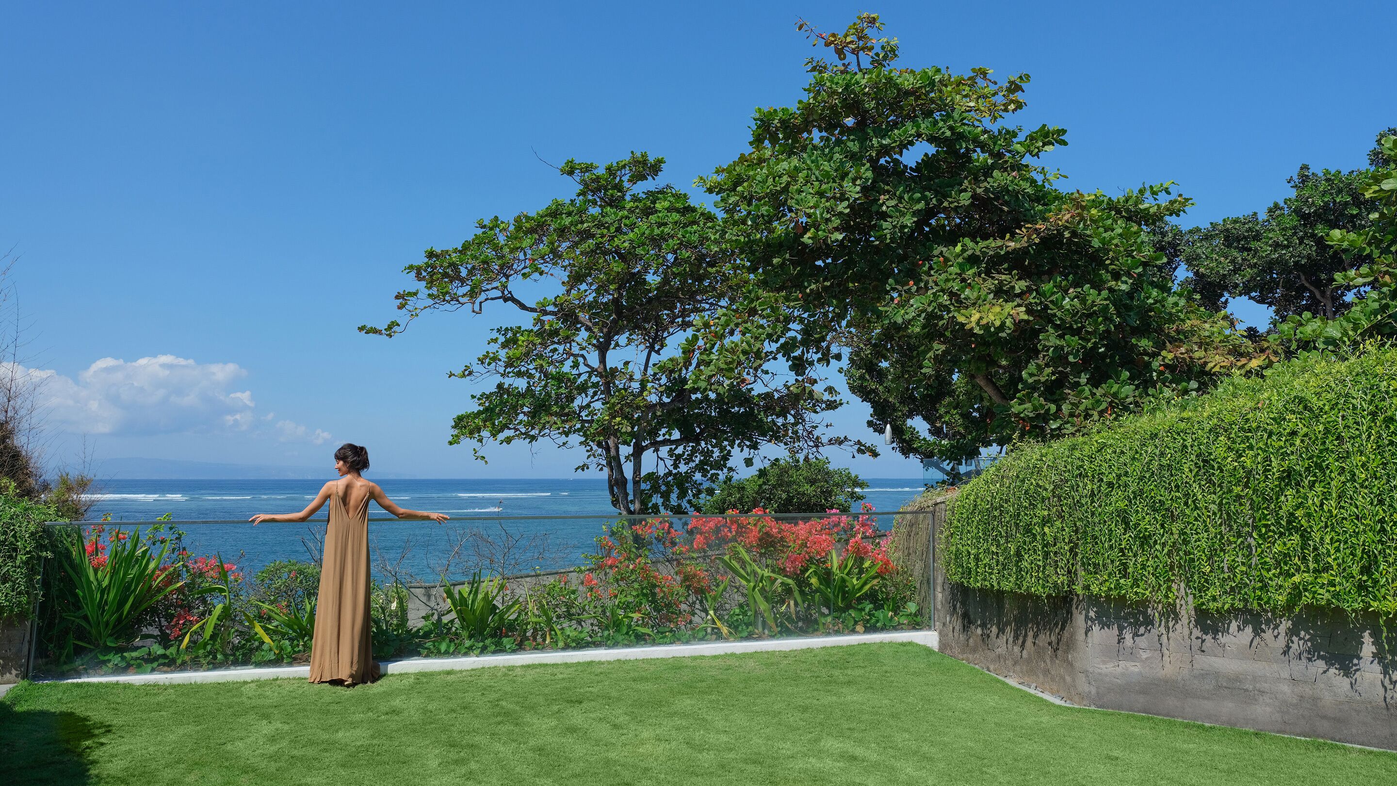 A woman in a long dress standing on a lawn with a view of the ocean and a tree.
