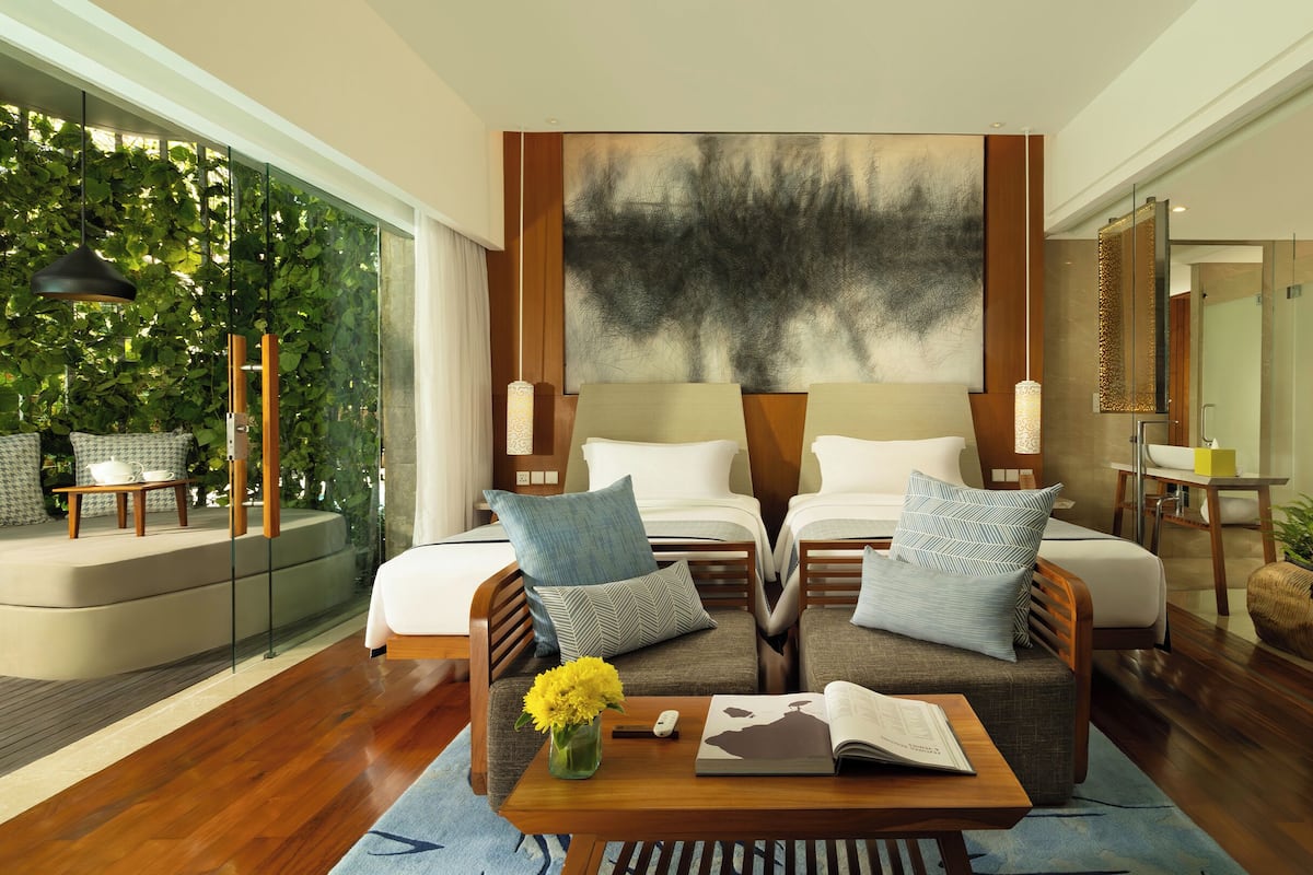 A modern hotel room with a large bed, two bedside tables, a sofa, a coffee table with a book and flowers, and a large window with a view of greenery.