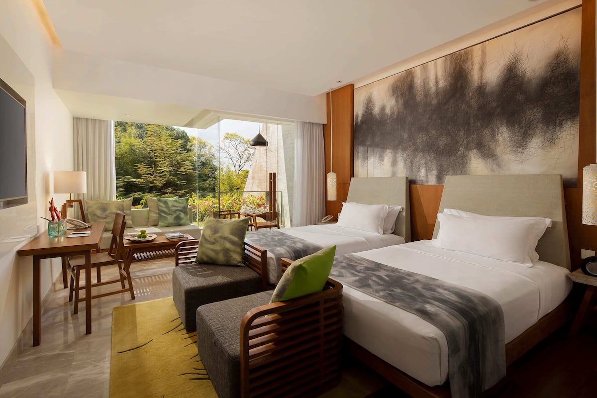 A hotel room with two beds, a desk, a chair, and a view of greenery.