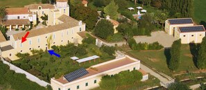 Eine Luftaufnahme eines Gebäudekomplexes mit mehreren Bauten, darunter ein großes zentrales Gebäude mit einem Ziegeldach, kleinere angrenzende Gebäude und Solaranlagen auf den Dächern.