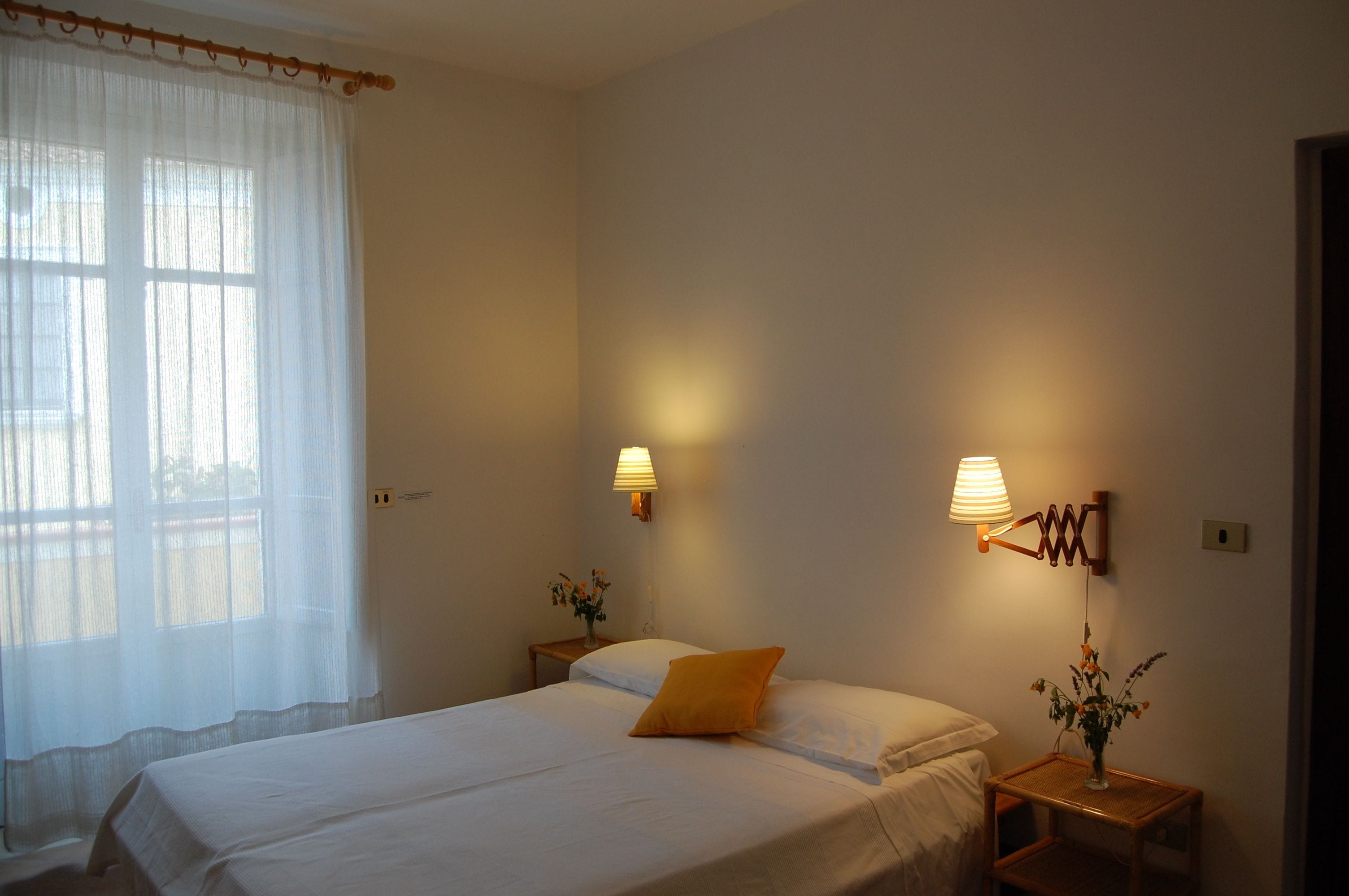 Un dormitorio con una cama, dos mesitas de noche, una lámpara y una ventana con cortinas traslúcidas.