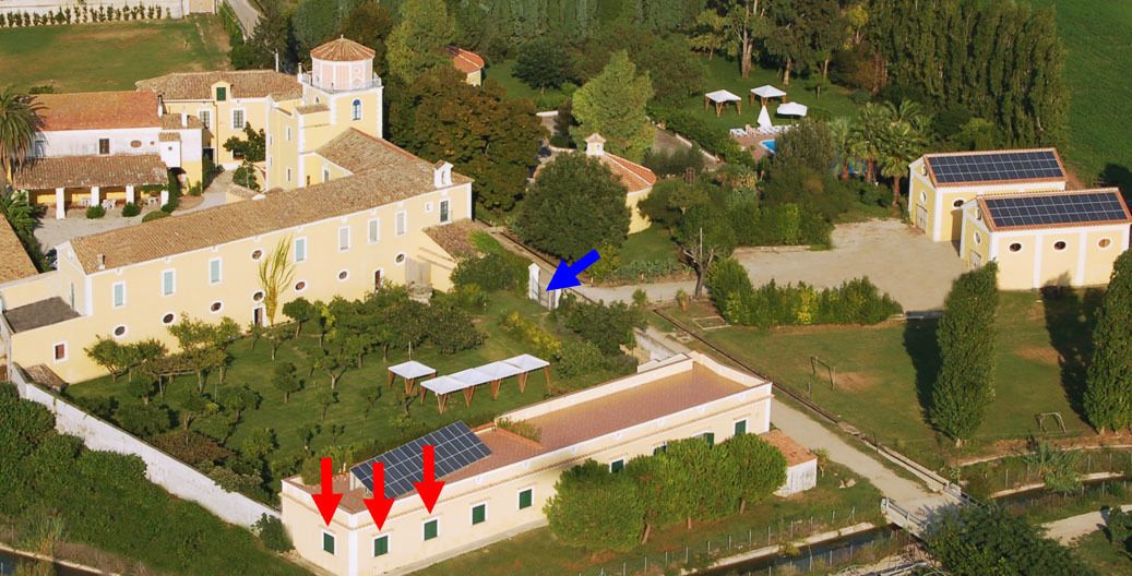 Vista aérea de un complejo histórico de edificios con varias construcciones, incluyendo un edificio central grande con una torre, varios edificios más pequeños y un patio con árboles y paneles solares.