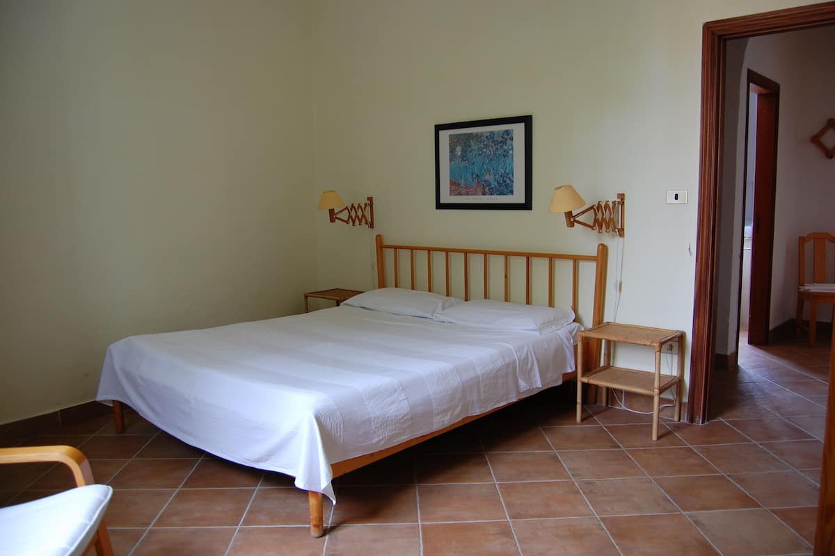 Une chambre à coucher avec un lit en bois, deux tables de chevet, une petite lampe de chevet, un tableau encadré au mur et une chaise.