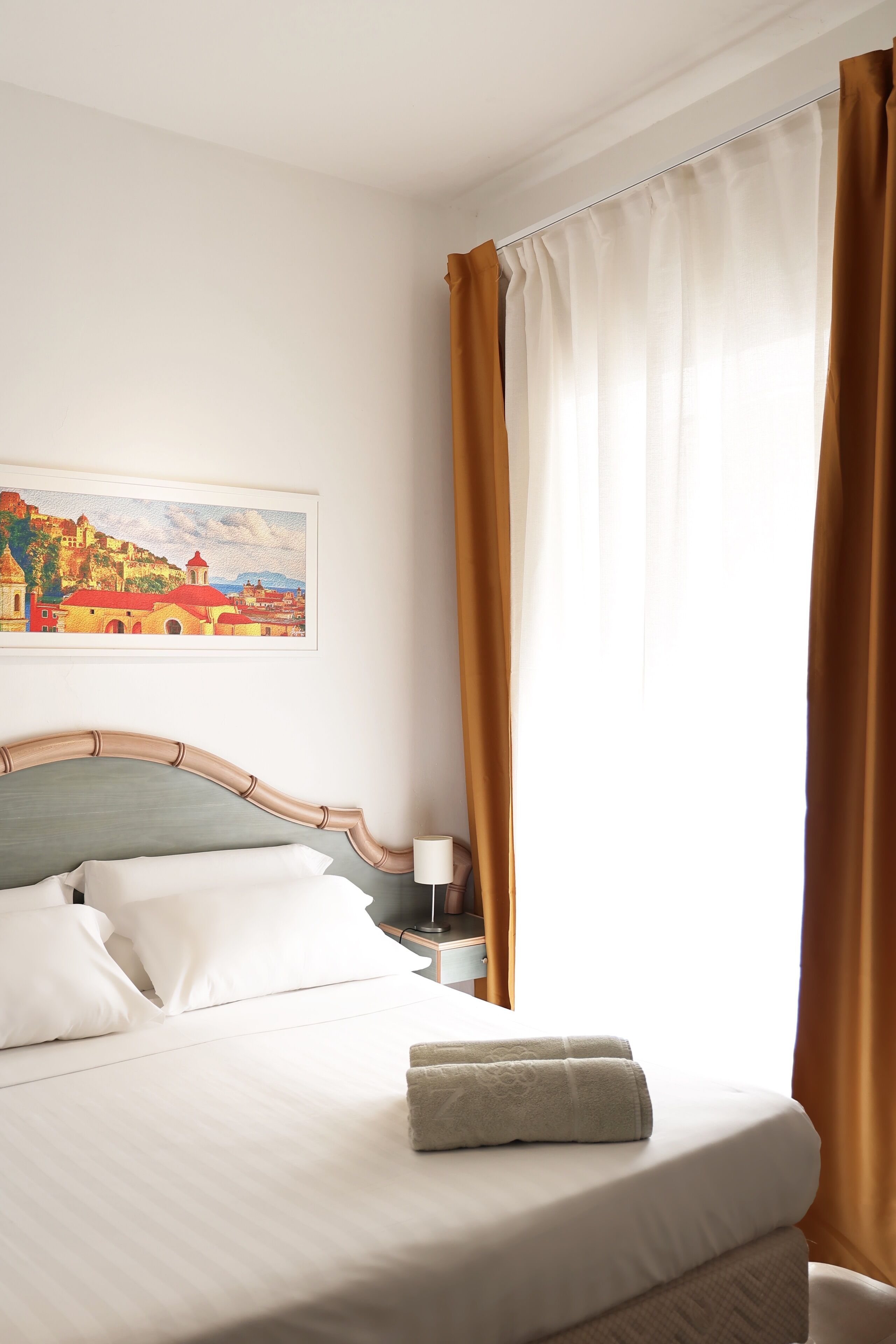 A neatly made bed with white linens and a folded towel, a bedside lamp, and a painting of a landscape on the wall.