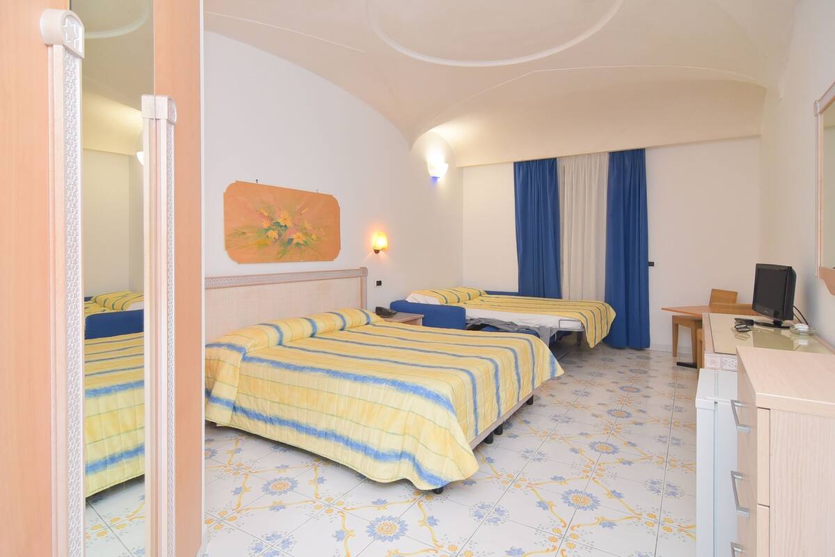 Une chambre d’hôtel avec trois lits, un bureau, une chaise, une télévision et un tableau au mur.