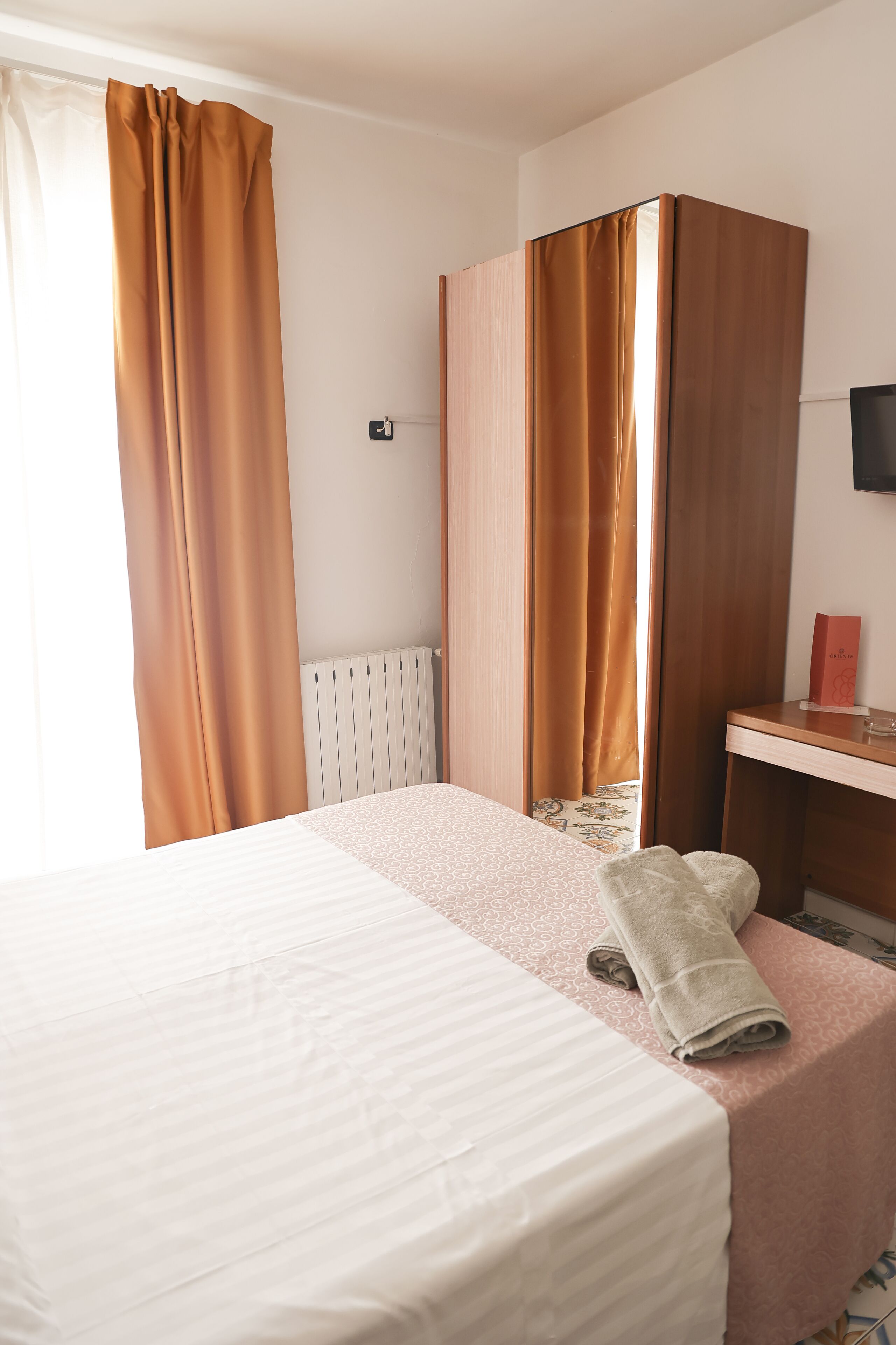 A hotel room with a bed, a wooden wardrobe, a desk with a television, and a window with curtains.