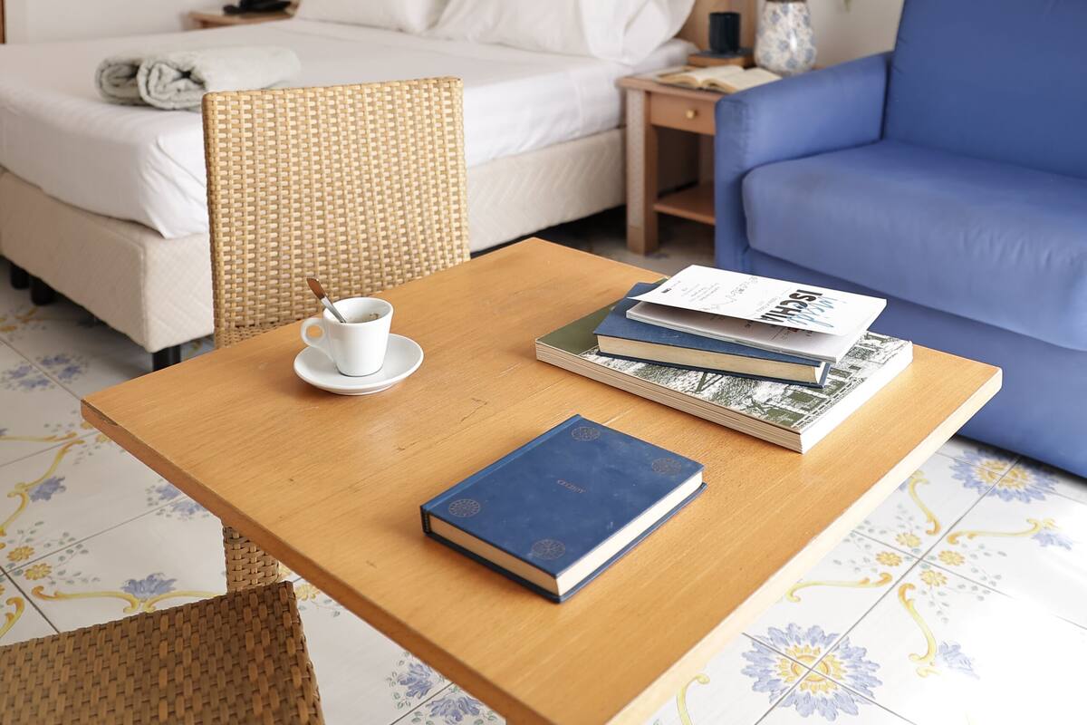 Une chambre d’hôtel avec un lit, un canapé bleu, une table en bois avec des livres et une tasse, et un tableau accroché au mur.