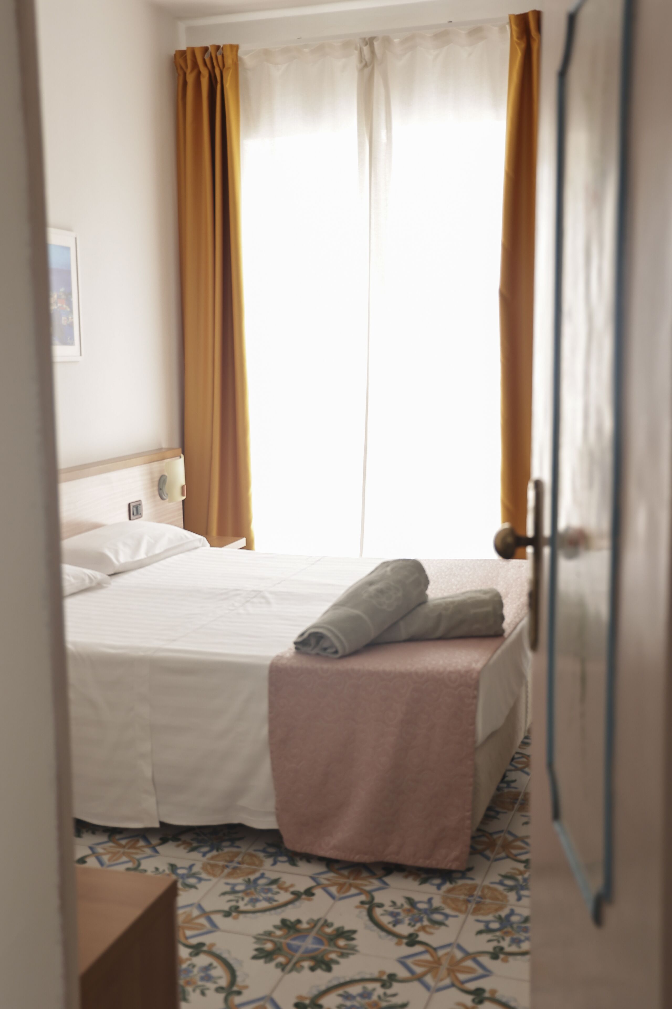 A hotel room with a bed, curtains, a bedside table, and a tiled floor.