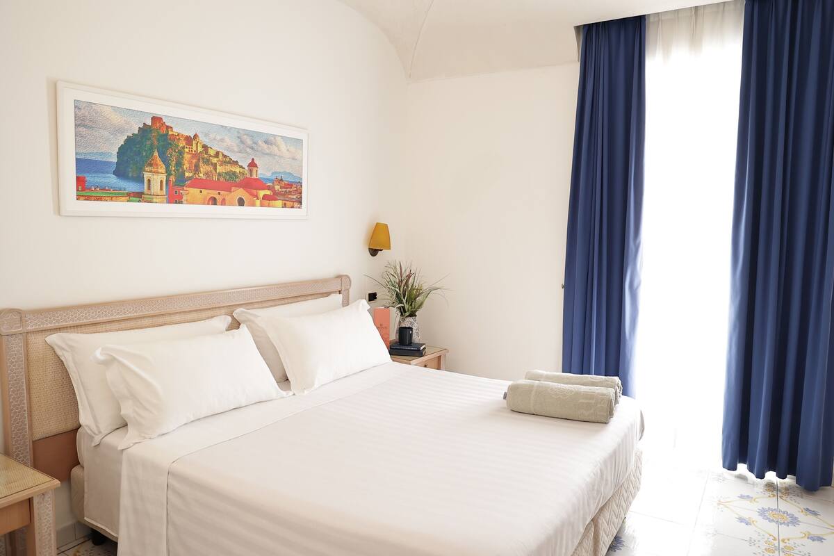 Un lit bien fait, avec du linge de lit blanc, un tableau encadré représentant un paysage côtier accroché au mur, et une table de chevet avec une lampe et une petite plante.