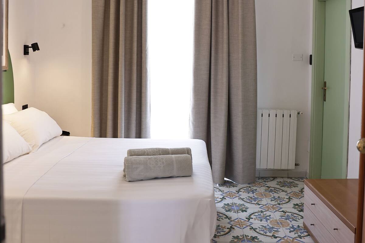Une chambre à coucher avec un lit, des rideaux, un radiateur, une commode et un téléviseur.