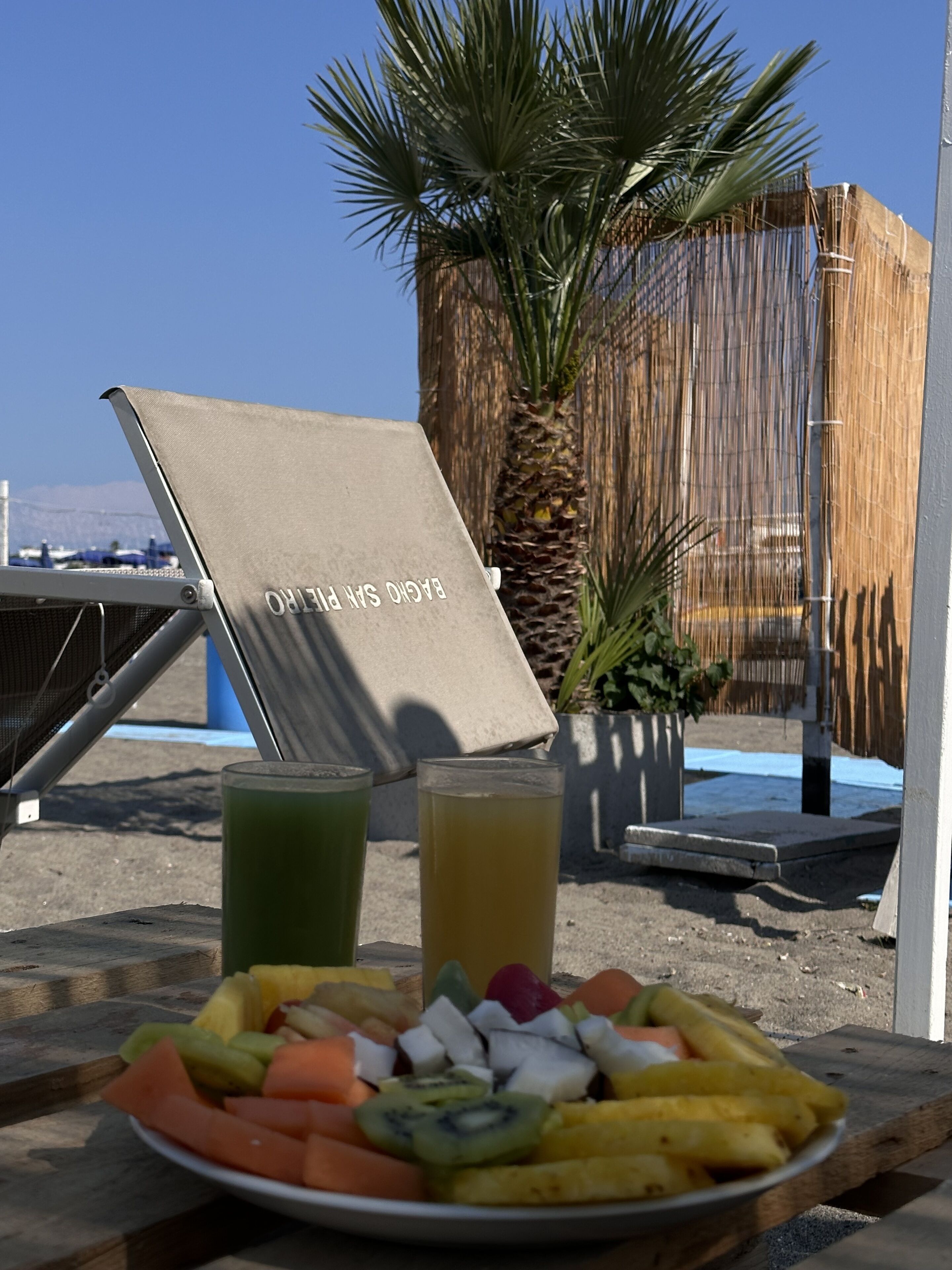 A table with a plate of fruit and two drinks, a beach umbrella, and a palm tree in the background.