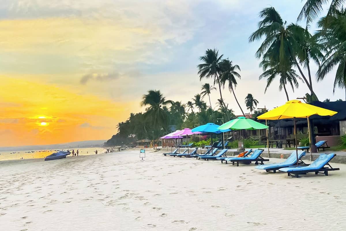 A beach with sun loungers and umbrellas, palm trees, and a sunset.