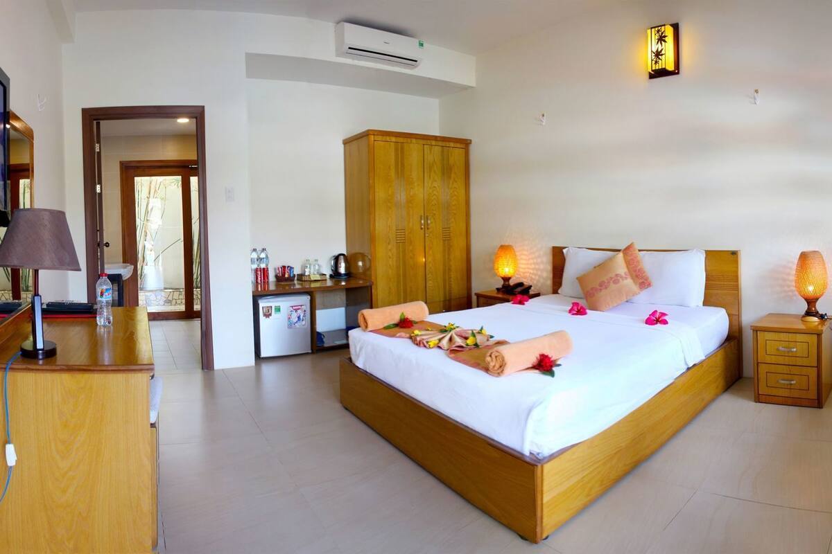 A hotel room with a bed, bedside tables, a desk, a wardrobe, a TV, and an air conditioning unit.