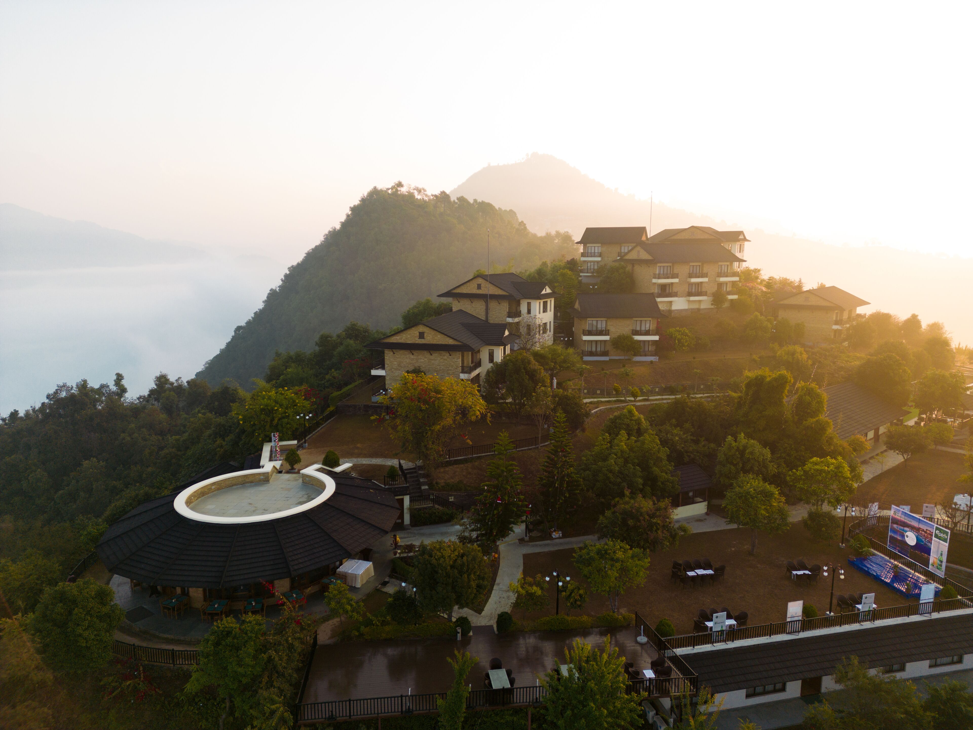 A hillside residential area with multiple houses, a circular building with a roof, and a view of a mountain in the background.