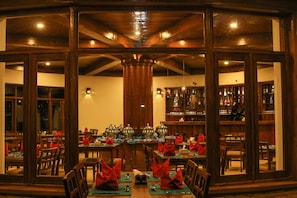 A restaurant with a long table set for a meal, wooden walls, and a bar area visible through the windows.