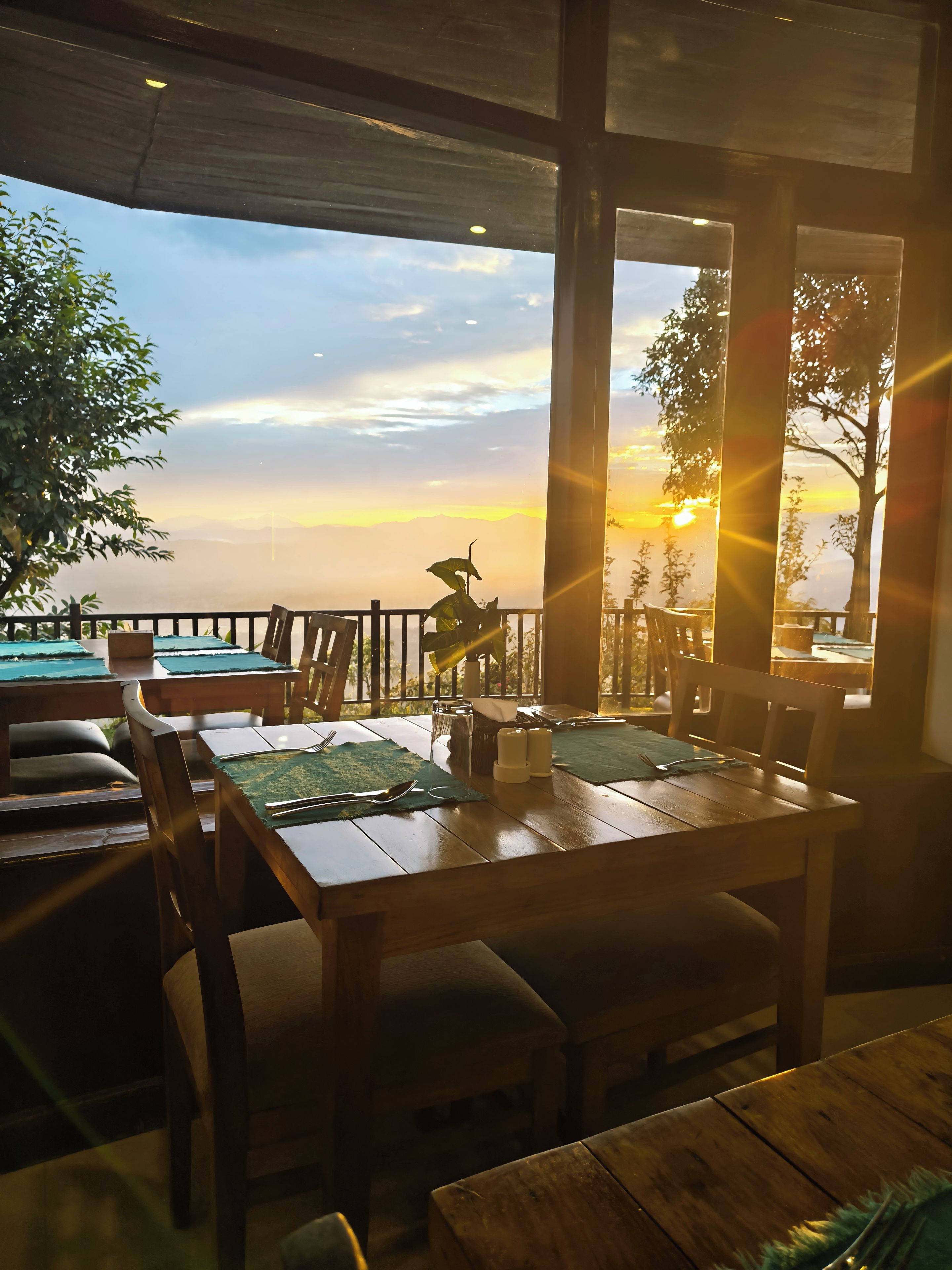 Un restaurante con mesas y sillas de madera, vista al atardecer y barandilla con vista al océano.