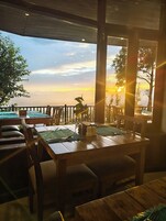 Un restaurante con mesas y sillas de madera, vista al atardecer y barandilla con vista al océano.