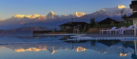 A pool with a mountain backdrop, lounge chairs, and a building with a roof.