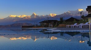 A pool with a mountain backdrop, lounge chairs, and a building with a roof.