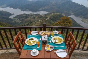 A table set with breakfast on a balcony overlooking a valley with a lake.
