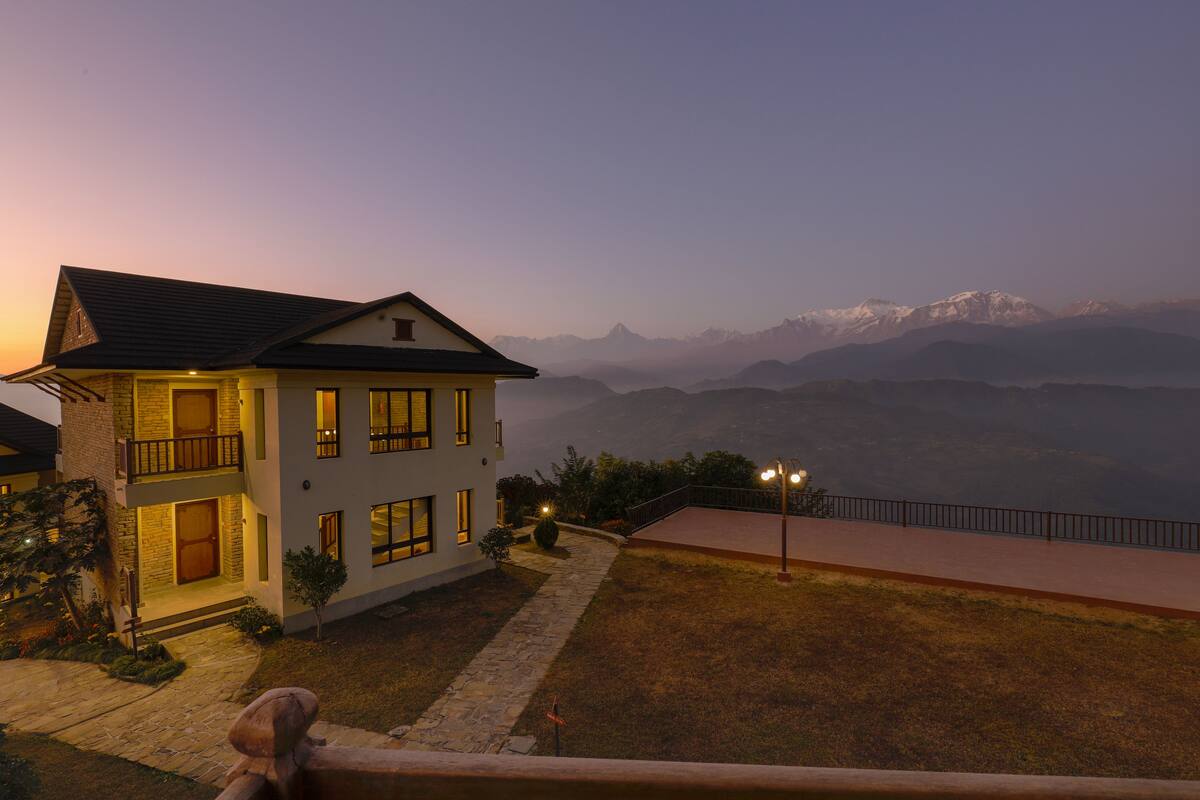 A two-story house with a balcony, a stone pathway, and a view of mountains in the background.