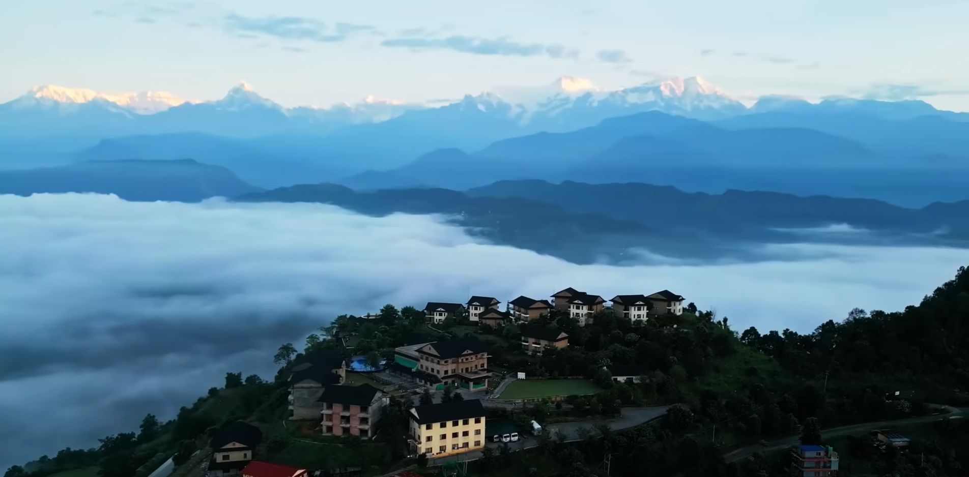 A mountain village with houses on a hillside, surrounded by clouds and snow-capped mountains in the background.