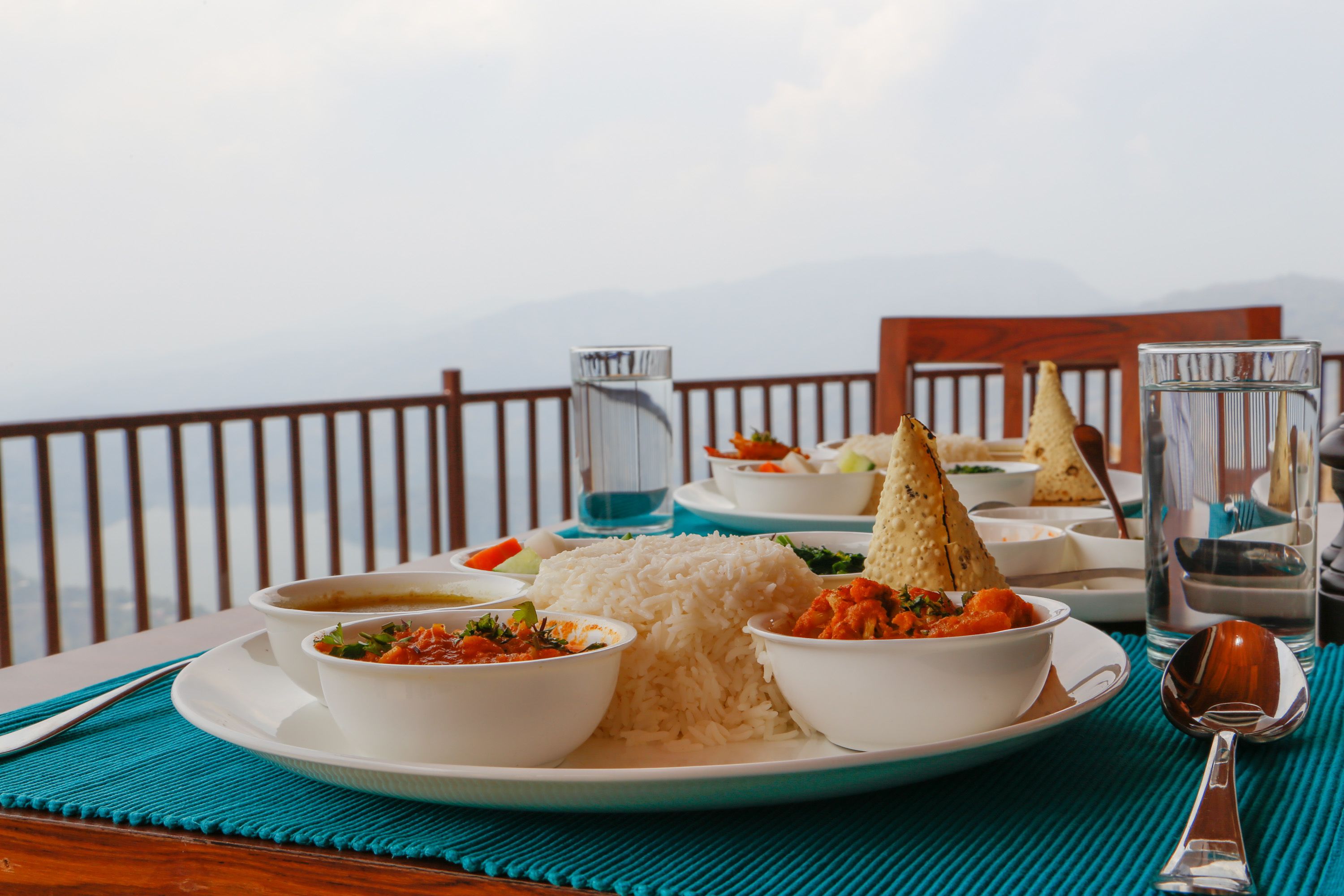 A table set with a plate of rice, two bowls of soup, and a glass of water, with a mountain view in the background.