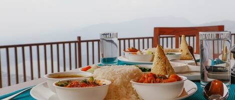 A table set with a plate of rice, two bowls of soup, and a glass of water, with a mountain view in the background.