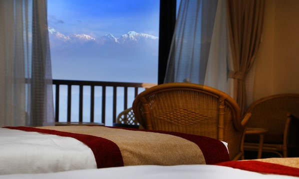 A hotel room with a bed, a wicker chair, and a view of snow-capped mountains through large windows.