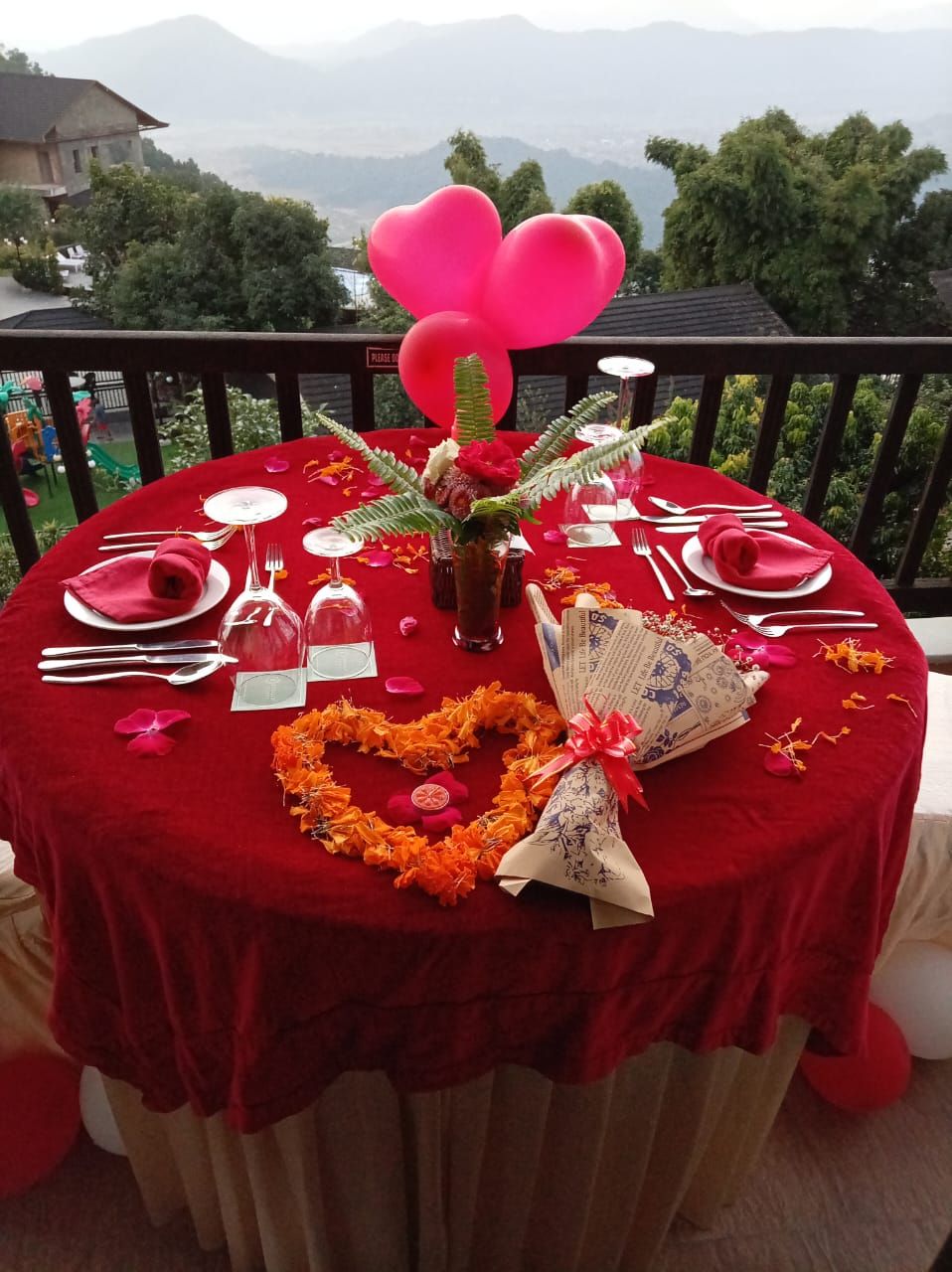 Une table ronde recouverte d’une nappe rouge, mise avec des assiettes blanches, des couverts en argent et un verre d’eau.