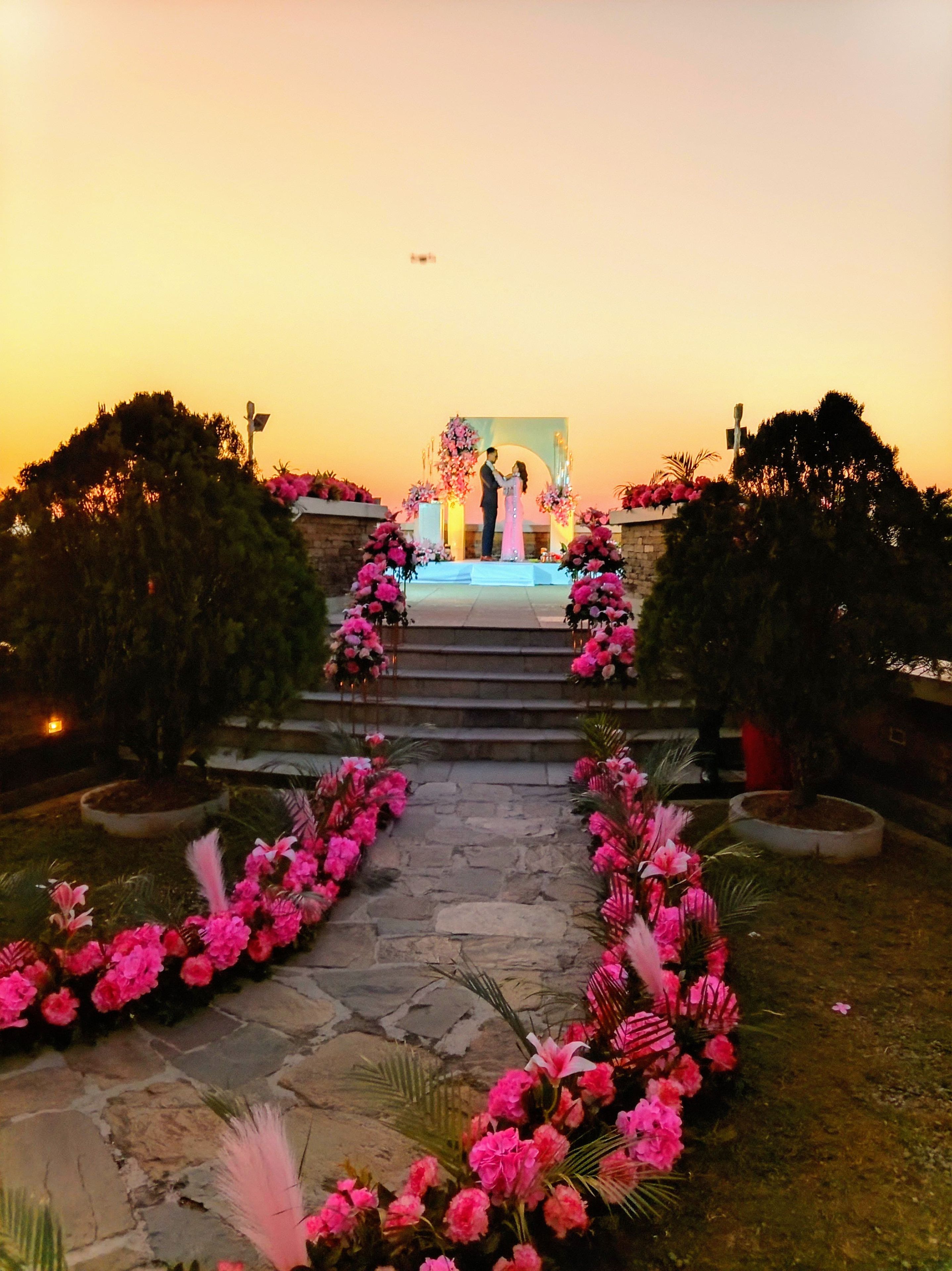 A stone pathway leading to a stage with a floral archway, surrounded by greenery.
