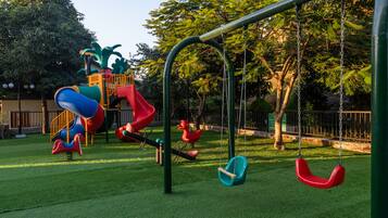 Un parque infantil con toboganes y columpios, rodeado de árboles y una valla.