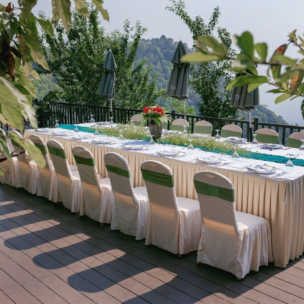 An outdoor dining area with a long table set for a meal, surrounded by chairs with green sashes, and a view of a forested area in the background.