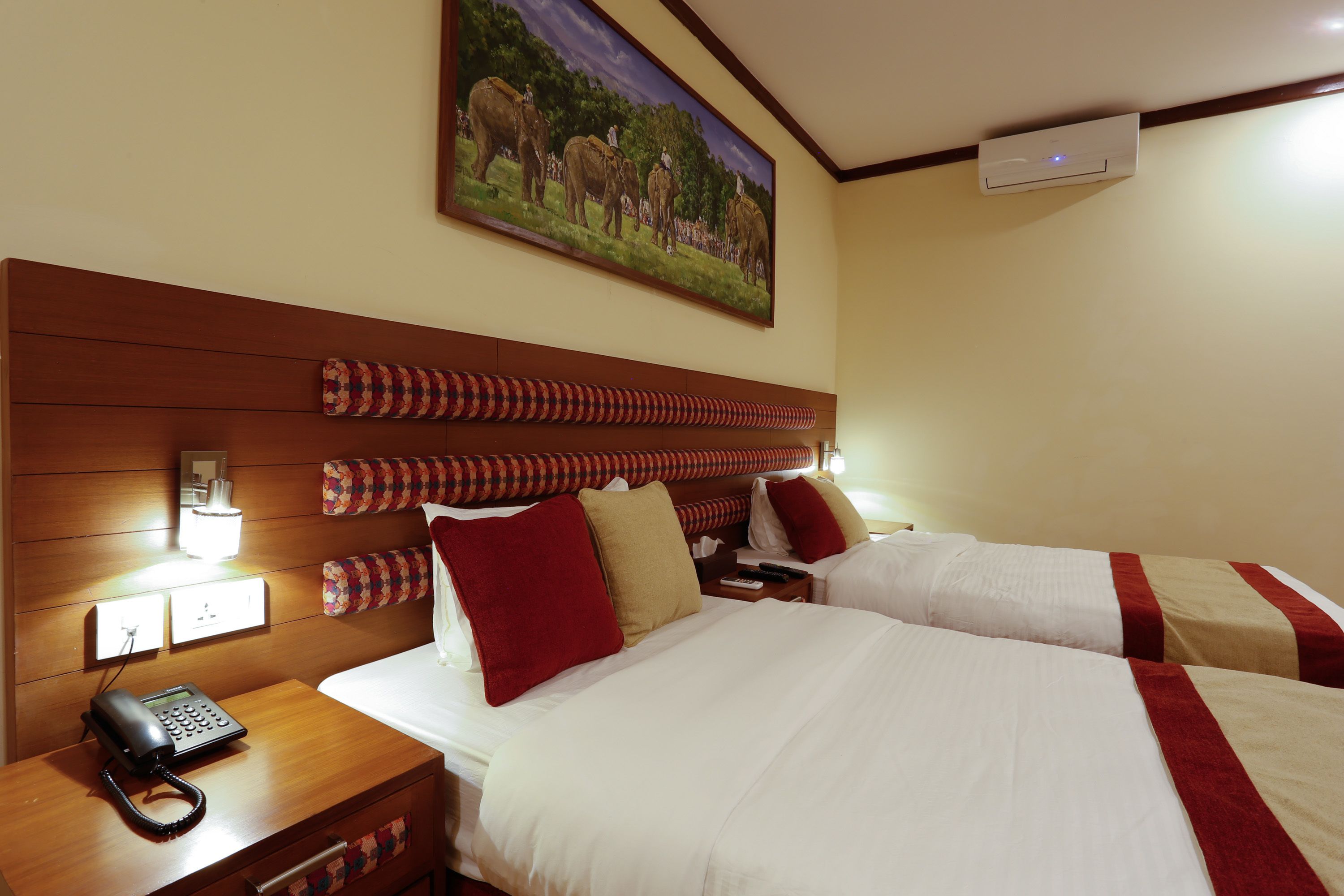 Une chambre d’hôtel avec deux lits, une tête de lit en bois, un tableau représentant des éléphants, un téléphone et une table de chevet.