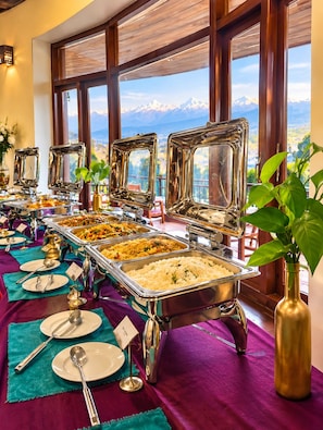 A buffet setup with multiple food stations, a view of mountains through large windows, and a purple tablecloth. - Rupakot Resort (Lekhnath)