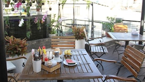 Une terrasse extérieure avec des tables et des bancs en bois, entourée de plantes en pot et de fleurs suspendues.