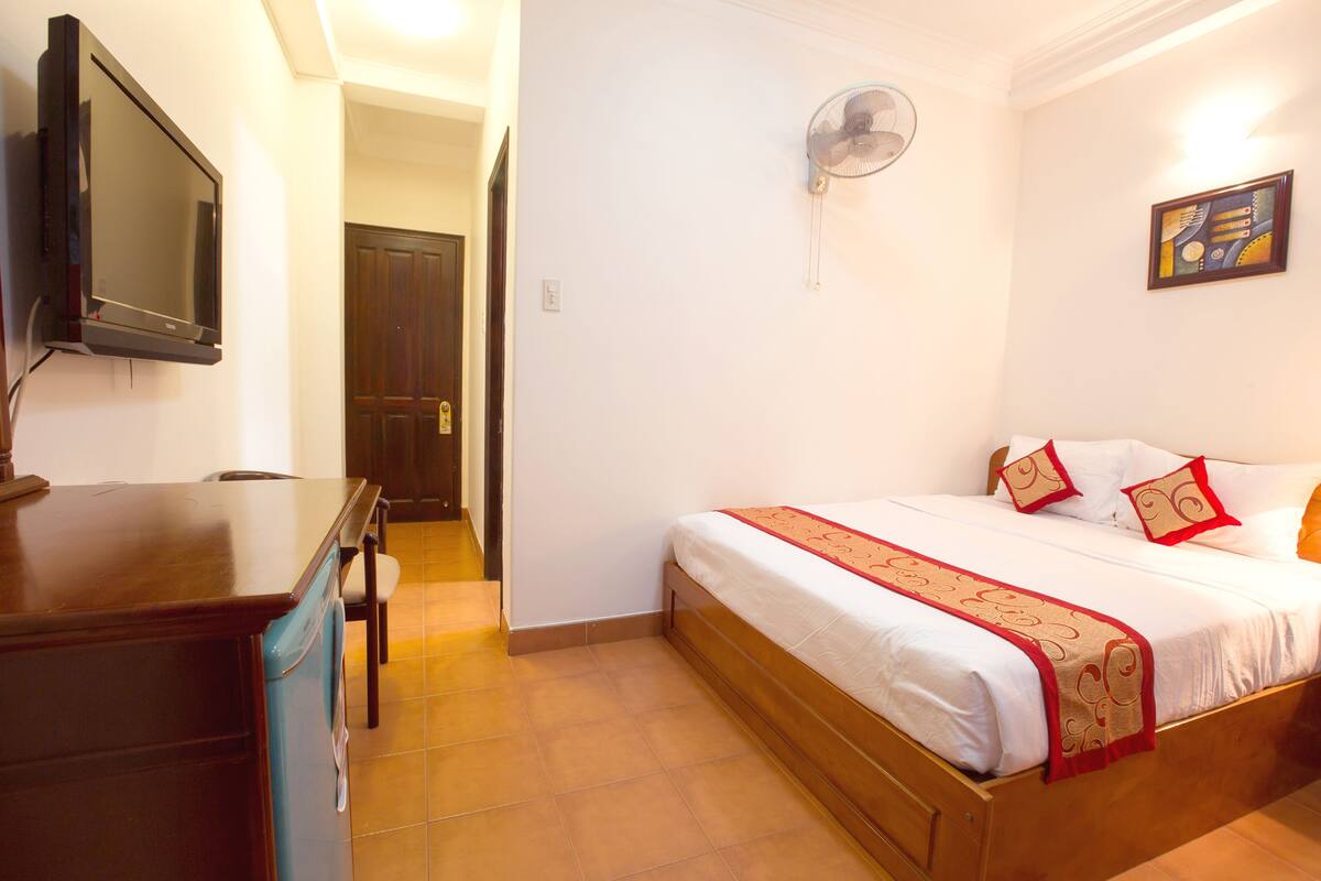 A bedroom with a bed, a desk, a chair, a television, a fan, and a door.