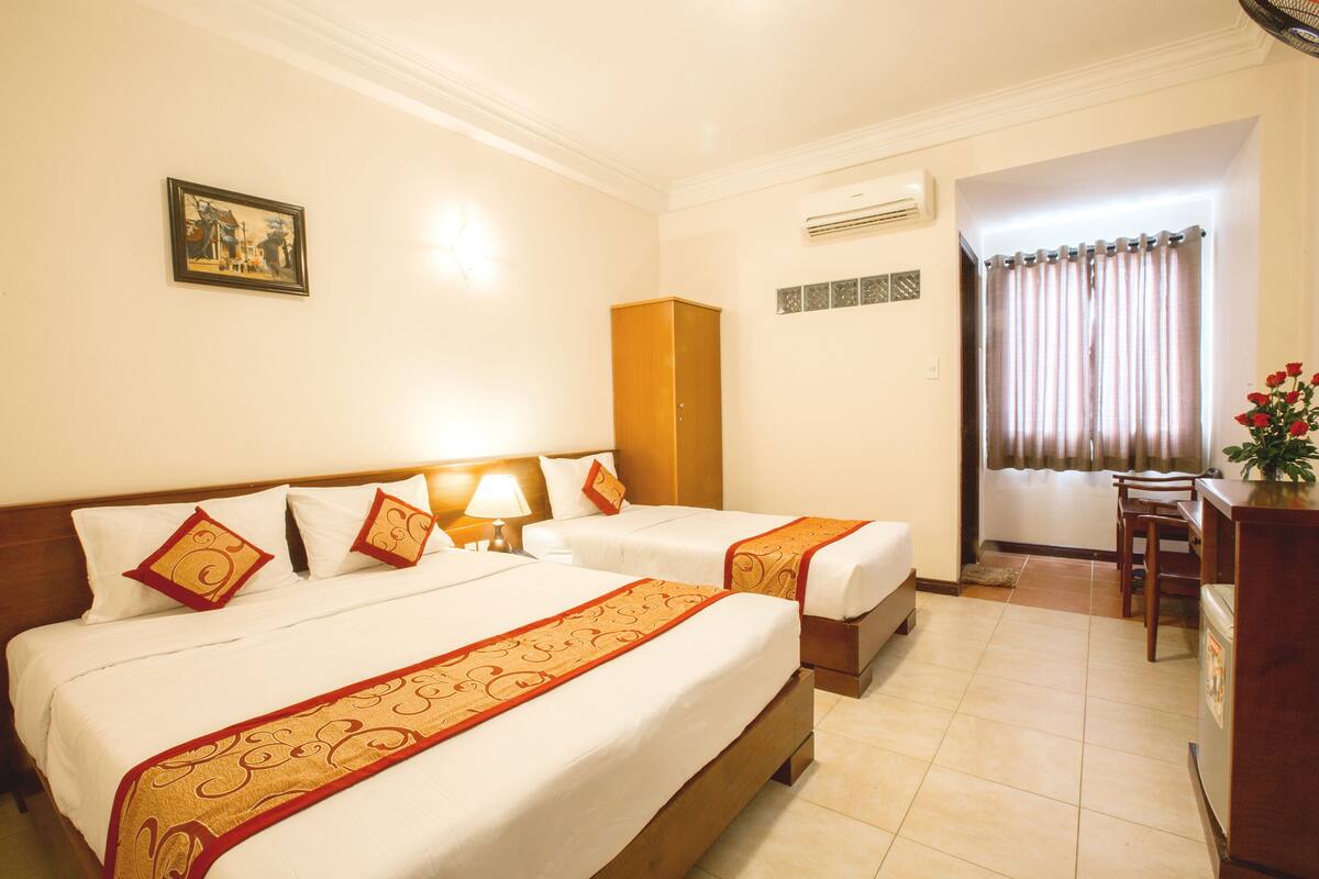 A hotel room with two beds, a wooden wardrobe, a desk with a chair, and a window with curtains.