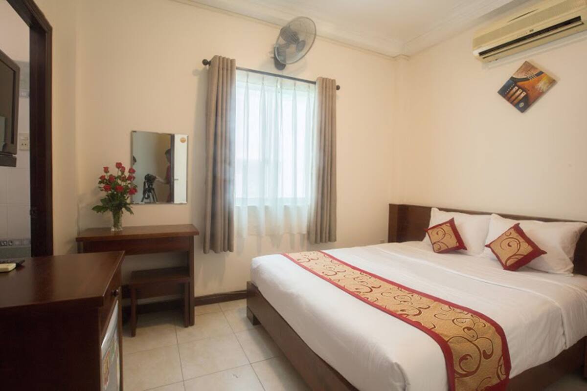 A hotel room with a bed, a desk, a mirror, a window with curtains, and an air conditioner.