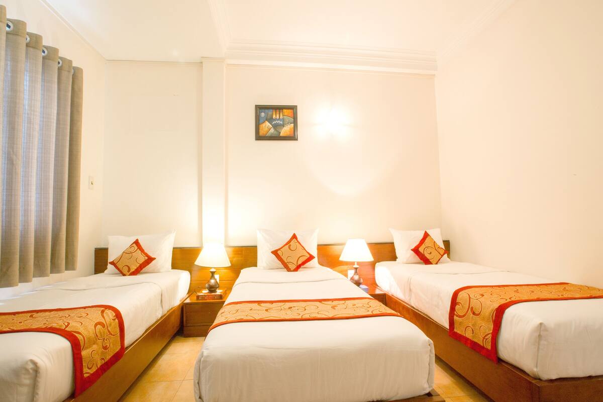 A hotel room with three single beds, each with a white bedspread and orange decorative trim. There are two bedside tables with lamps, and a small painting on the wall.