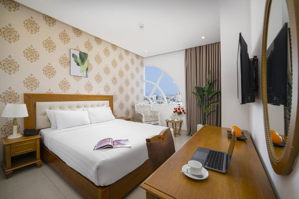 A hotel room with a large bed, a desk with a laptop, a cup, and a view of a cityscape through a window.