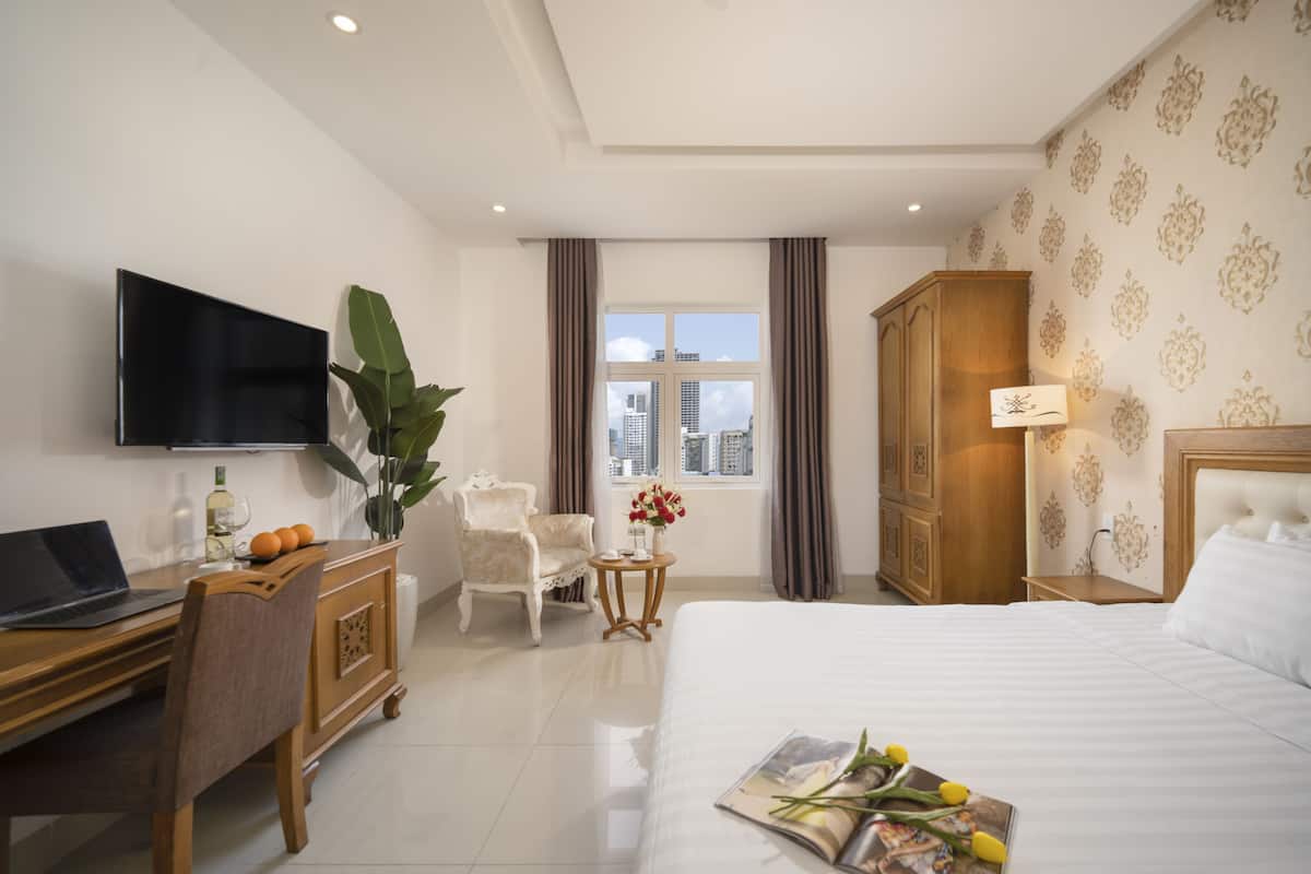 A hotel room with a bed, a desk with a laptop, a chair, a TV, a window with curtains, and a view of buildings.