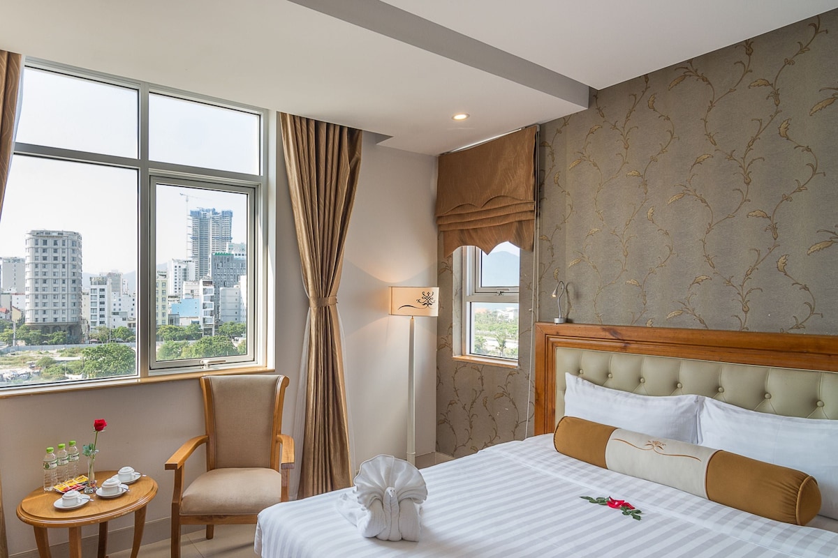 A hotel room with a bed, a chair, a small table, and a view of the cityscape through large windows.