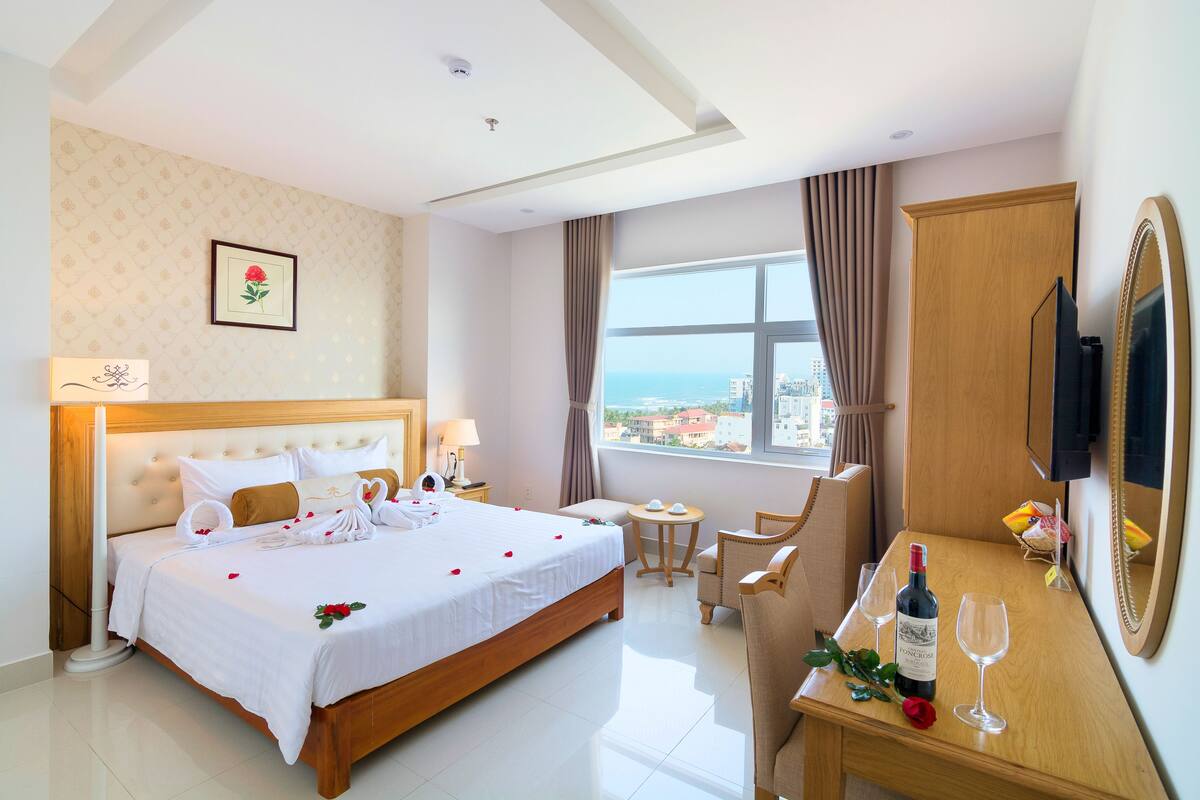 A hotel room with a large bed, a small table with a bottle and glasses, a chair, a TV, and a window with a view of the city.