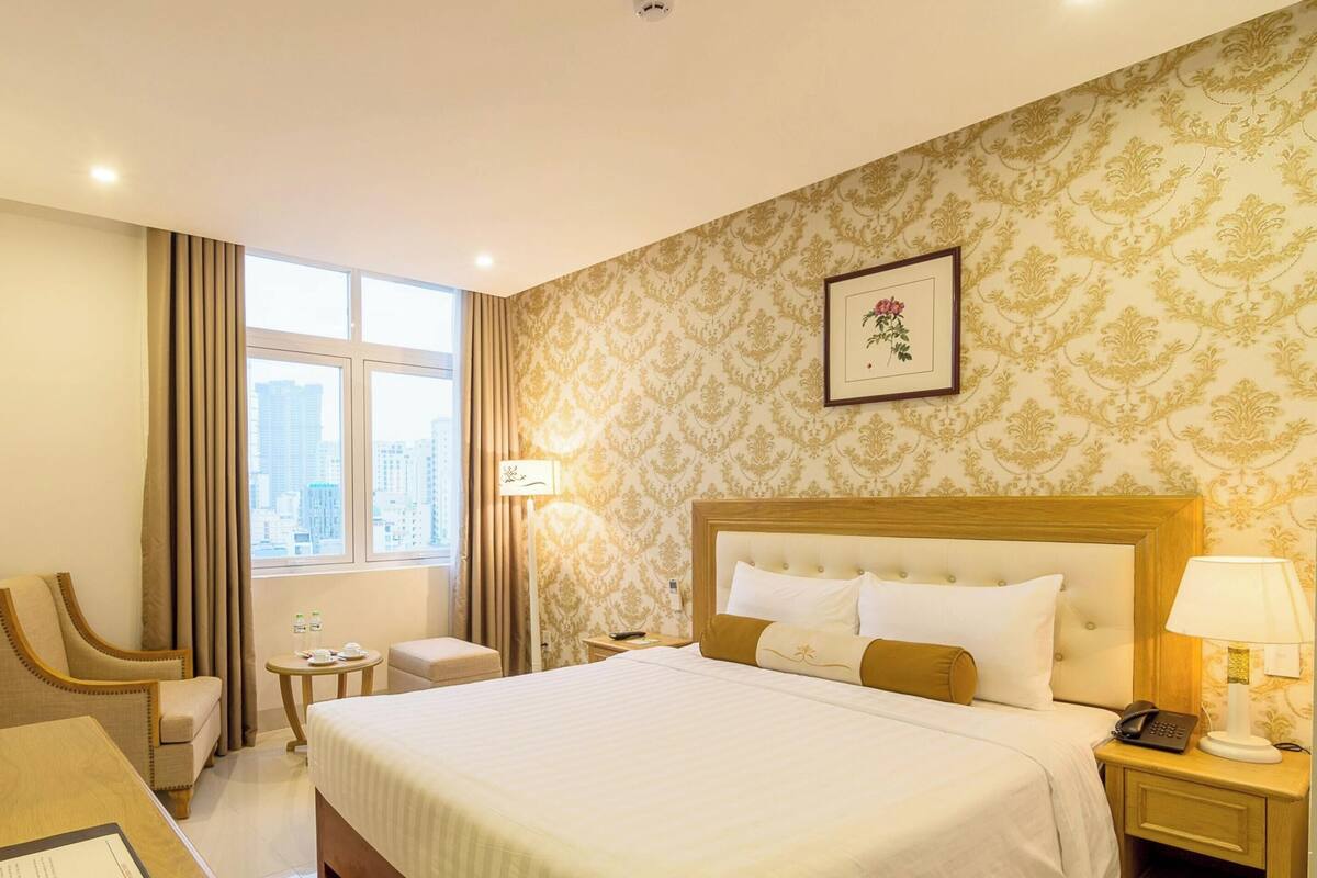 A hotel room with a bed, a desk, a chair, a bedside table, a lamp, and a window with a city view.