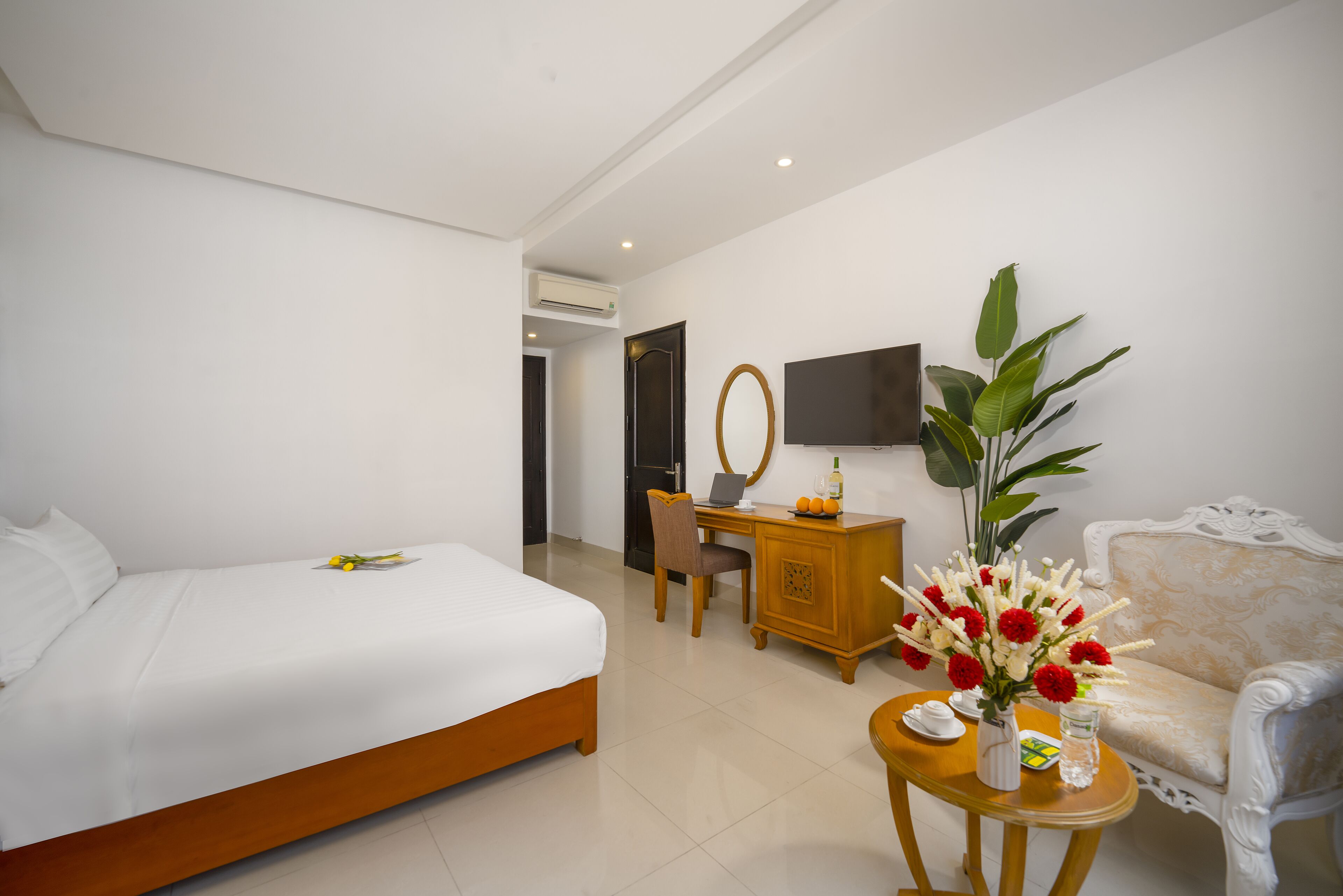 A hotel room with a bed, a desk, a chair, a TV, a sofa, and a table with flowers.