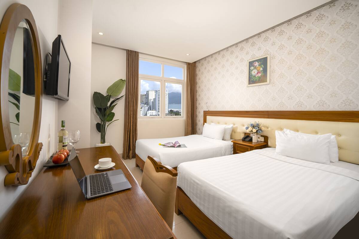 A hotel room with two beds, a desk with a laptop, a mirror, a window with a city view, and a decorative wall piece.