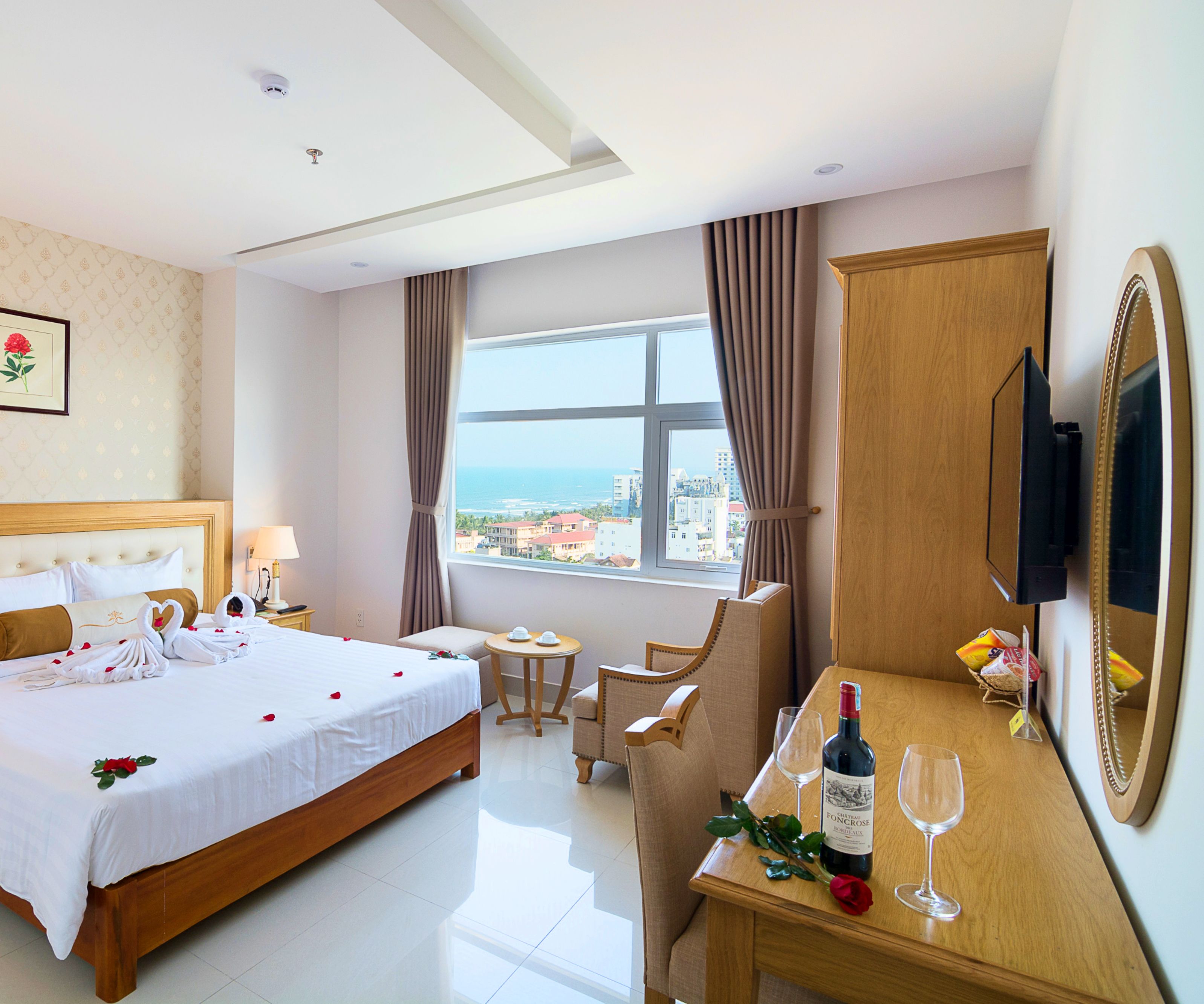 Une chambre d’hôtel avec un lit, un bureau sur lequel se trouve une bouteille de vin et des verres, une chaise, une télévision et une fenêtre donnant sur des immeubles et la mer.